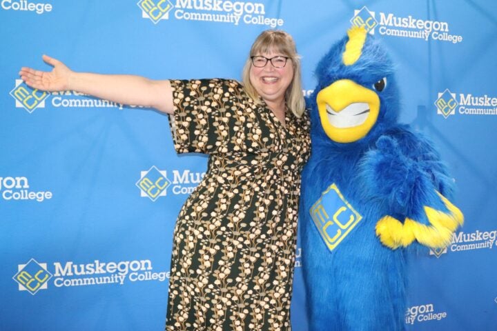 A woman in a gold and black dress stretches her arm out to the left and hugs MCC's Mascot, Jay the Jayhawk, a bird costume that is over five feet tall. Behind them is a wall displaying MCC logos.