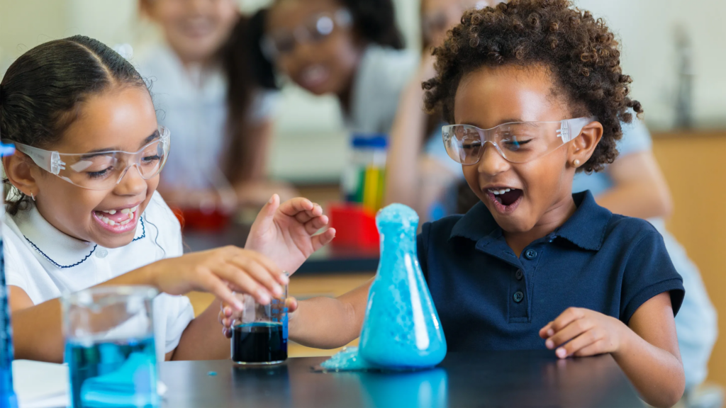 Two young children laugh in wonder as they conduct a science experiment while wearing safety goggles.