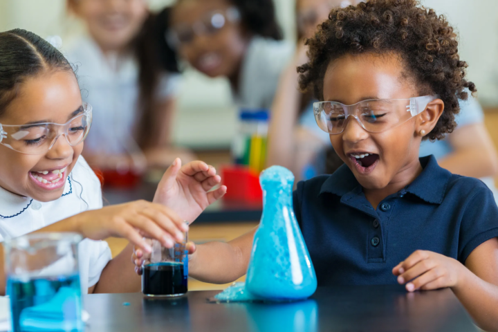 Two young children laugh in wonder as they conduct a science experiment while wearing safety goggles.