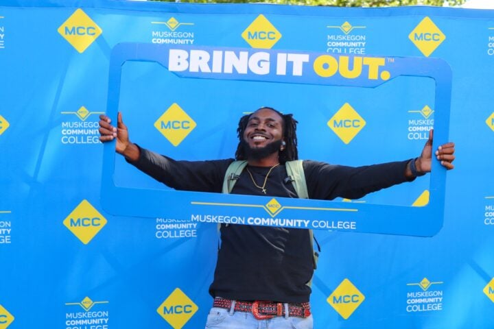 A student smiles in front of a blue MCC backdrop and holds up a life size license plate frame as he poses inside for the camera.