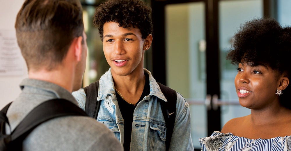 Two african american students talk to a man with a backpack on in the hallway of campus.
