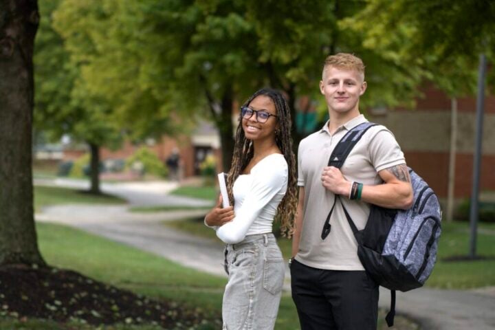 A White High School Student and an African-American female high student smile for the camera and hold backpacks outside.