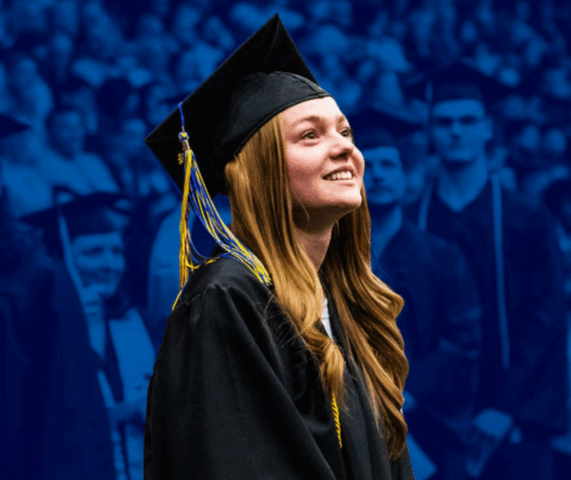 Girl smiles in front of a crowd. She is wearing graduation attire and looking upwards.