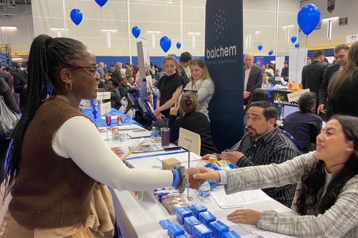 A girl shakes hands with someone seated at a table. There are many people in the background. It is a jobs fair and there are balloons floating up