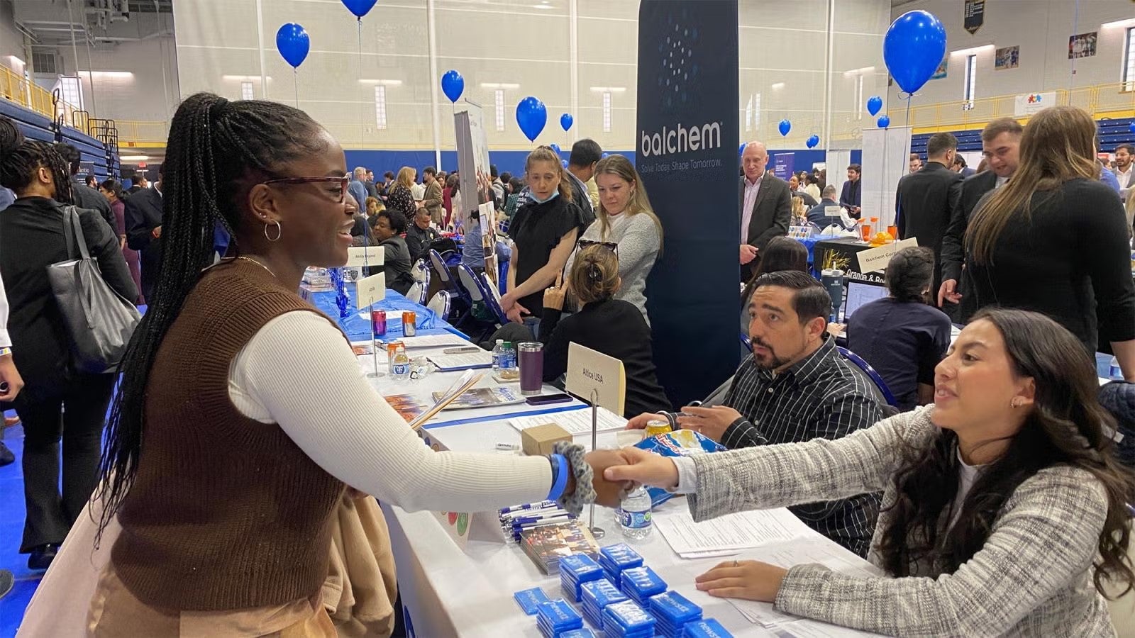 A girl shakes hands with someone seated at a table. There are many people in the background. It is a jobs fair and there are balloons floating up
